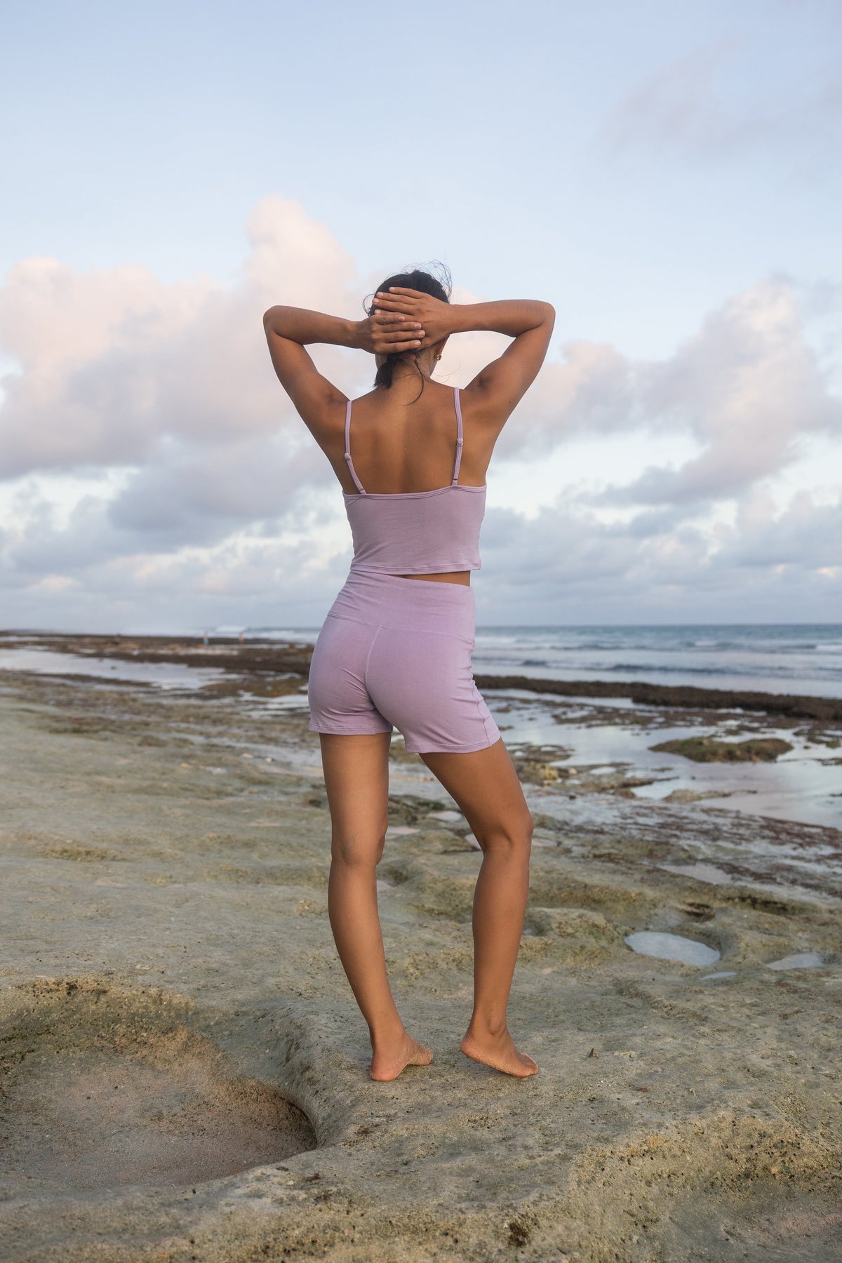 Woman standing on a tide pool beach wearing an Adjustable Cotton Cami and matching shorts in Lilac, back view, hands behind head #Color_Lilac